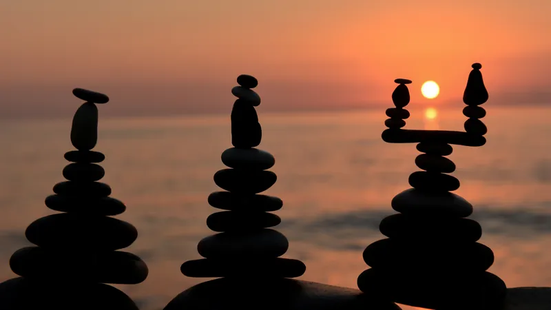 Three balanced stone cairns silhouetted against a burning sunset over still water, the balance between an outer life and an inner one
